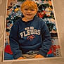 blonde_hair, blue_clothing, child, christmas_tree, decoration, fabric_surface, festive, happy, holiday, home, indoor, ornaments, person, photo_print, portrait, red_decor, seasonal, smiling, sweatshirt, winter