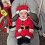 baby, santa_claus_outfit, red_clothing, baby_chair, toy, holiday, christmas, festive, gift_box, carpet, indoor, smiling, cute, child, face, hat, black_boots, seatbelt, cushion, decorations