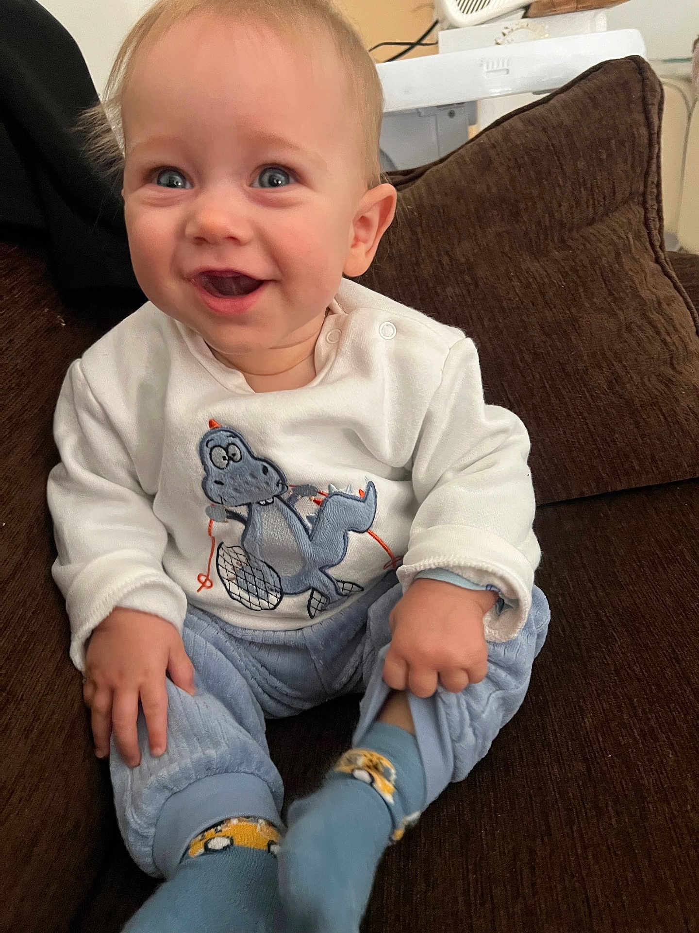 Lyham a rejoint le concours — aidez-le/la à gagner de superbes lots ! baby, child, smiling, blue_eyes, white_shirt, dinosaur_graphic, blue_pants, socks, sitting, couch, indoor, happy, cute, infant, portrait, hands, feet, young_child, cozy, casual_clothing