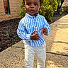 child, toddler, boy, portrait, standing, outdoor, sidewalk, striped_shirt, blue_shirt, white_pants, black_shoes, curly_hair, dark_skin, serious_expression, fashion, formalwear, building, garden, shrubs, daylight