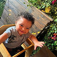 Jaylin joined the competition — help win amazing prizes! child, toddler, curly_hair, high_chair, smiling, reaching_hand, wooden_furniture, greenery, plants, flowers, string_lights, indoor, casual_clothing, happy, cute, portrait, baby, seat, face, person