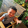 child, toddler, curly_hair, high_chair, smiling, reaching_hand, wooden_furniture, greenery, plants, flowers, string_lights, indoor, casual_clothing, happy, cute, portrait, baby, seat, face, person