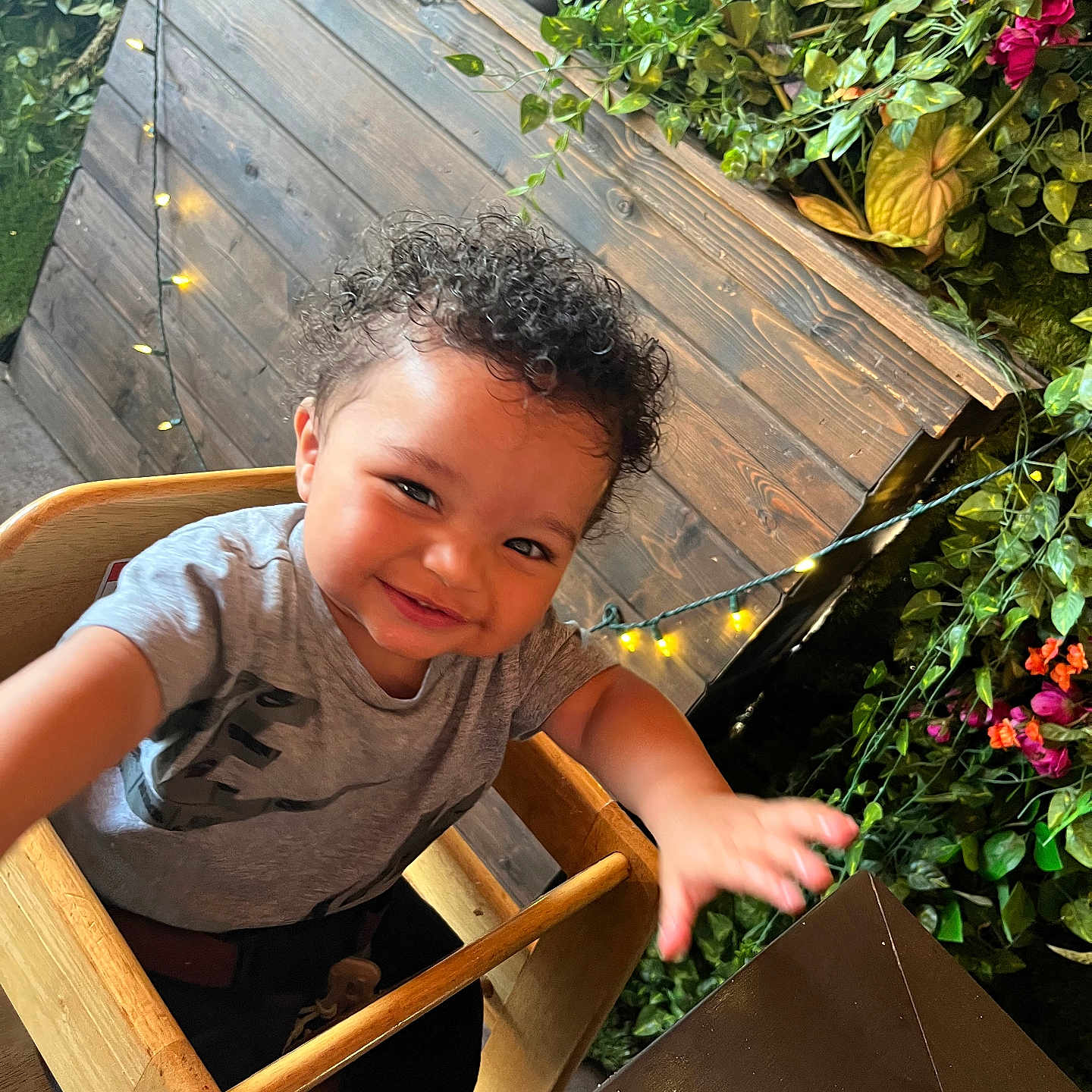 Jaylin joined the competition — help win amazing prizes! child, toddler, curly_hair, high_chair, smiling, reaching_hand, wooden_furniture, greenery, plants, flowers, string_lights, indoor, casual_clothing, happy, cute, portrait, baby, seat, face, person