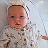 Nora is registered to the contest to win money with this photo: baby, infant, child, portrait, face, floral_outfit, dress, headband, bow, white_clothing, rosy_cheeks, eyes_open, looking_at_camera, lying_down, bed, blanket, indoor, cozy, close_up, photography