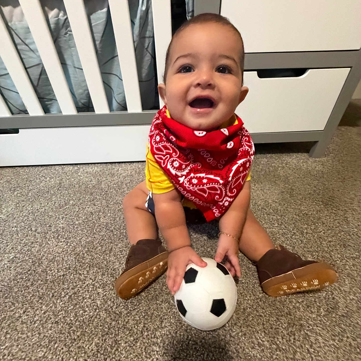 Maximilian is registered to the contest to win money with this photo: baby, child, infant, smiling, happy, sitting, soccer_ball, football, bandana, red_clothing, boots, carpet, nursery, crib, furniture, indoors, portrait, playtime, sports, cute