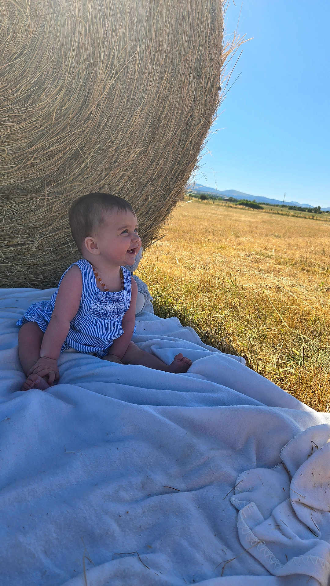 Naomie a rejoint le concours — aidez-le/la à gagner de superbes lots ! baby, child, blanket, hay_bale, outdoor, field, grass, sunlight, sky, blue_sky, smiling, sitting, summer, nature, portrait, happy, one_person, casual_clothing, daylight, rural