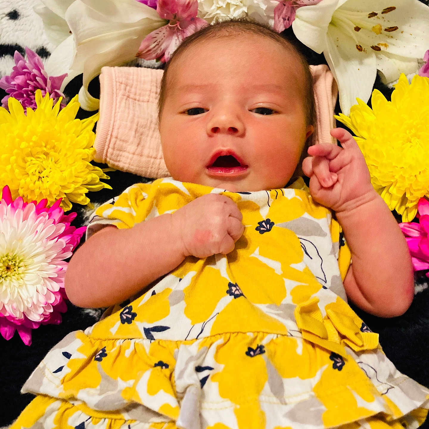Alora joined the competition — help win amazing prizes! baby, infant, flower, yellow_dress, floral_pattern, face, hand, lying_down, colorful, cute, portrait, newborn, soft_texture, pink_flower, white_flower, purple_flower, yellow_flower, black_background, closeup, peaceful