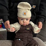 Jett joined the competition — help win amazing prizes! baby, child, beanie, hat, overalls, brown_clothing, cream_clothing, fire_truck_patch, couch, indoor, person, adult_hands, supporting, flooring, curious_expression, pouty_lips, seated, cute, clothing_brand, closeup