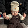 baby, child, beanie, hat, overalls, brown_clothing, cream_clothing, fire_truck_patch, couch, indoor, person, adult_hands, supporting, flooring, curious_expression, pouty_lips, seated, cute, clothing_brand, closeup