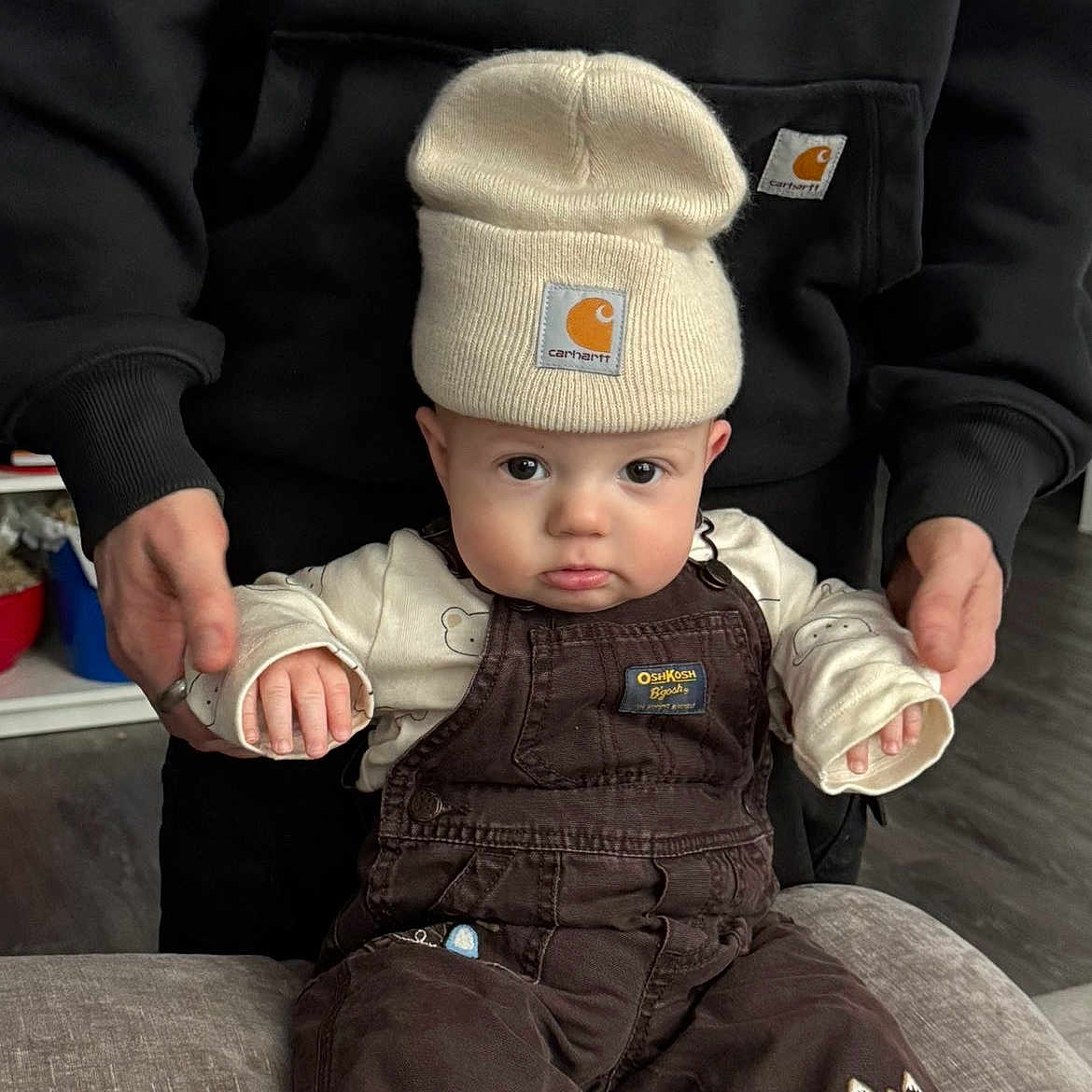 Jett joined the competition — help win amazing prizes! baby, child, beanie, hat, overalls, brown_clothing, cream_clothing, fire_truck_patch, couch, indoor, person, adult_hands, supporting, flooring, curious_expression, pouty_lips, seated, cute, clothing_brand, closeup