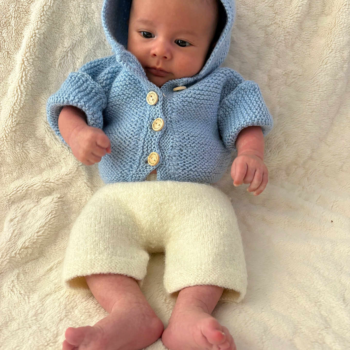 Aswan a rejoint le concours — aidez-le/la à gagner de superbes lots ! baby, bare_feet, blue_clothing, child, cozy, cream_pants, curious_expression, cute, fist, hoodie, indoors, infant, knitted_clothing, lying_down, newborn, portrait, relaxed, small_hands, soft_blanket, warm