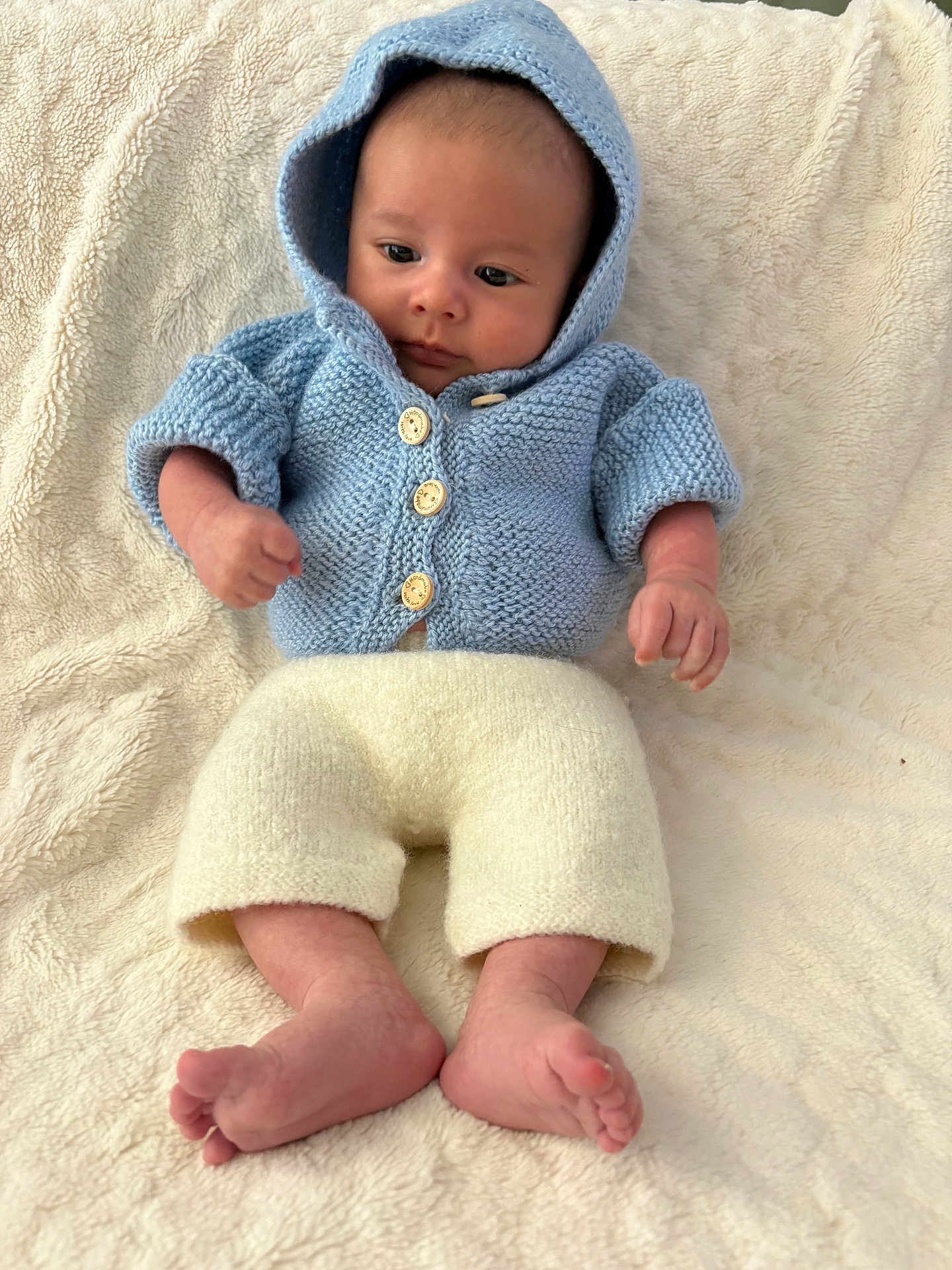 Aswan a rejoint le concours — aidez-le/la à gagner de superbes lots ! baby, infant, hoodie, knitted_clothing, blue_clothing, cream_pants, bare_feet, fist, curious_expression, soft_blanket, cozy, indoors, child, portrait, newborn, cute, warm, relaxed, lying_down, small_hands