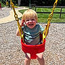 Xavier joined the competition — help win amazing prizes! child, toddler, swing, playground, yellow_chains, red_swing, smiling, happy, outdoor, summer, grass, wood_chips, sidewalk, trees, fence, greenery, sunlight, shorts, sandals, park