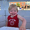 child, toddler, smiling, messy_face, red_tank_top, outdoor, table, bench, wooden_furniture, daylight, blue_sky, building, people, blond_hair, happy, cute, face, person, casual_clothing, summer