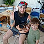 man, boy, dog, tattoos, cap, sunglasses, wooden_bench, indoor, plants, stroller, tile_floor, smiling, shorts, sleeveless_shirt, child, pet, casual, family, beard, home