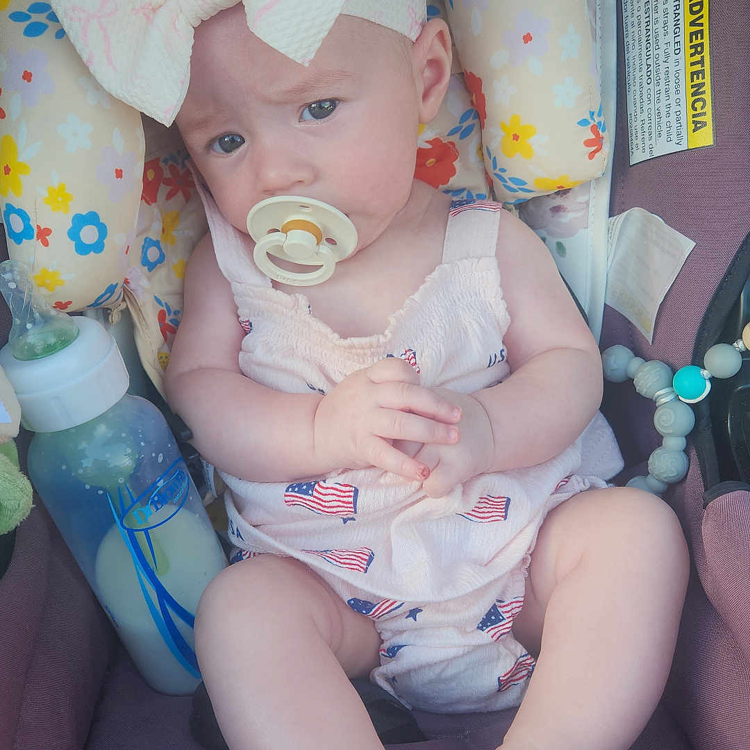 Olive joined the competition — help win amazing prizes! baby, child, infant, car_seat, bow, pacifier, pink_clothing, dress, sitting, portrait, face, smiling, eyes, hands, feet, bottle, baby_toy, floral_pattern, indoors, automobile