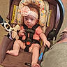 Olive is registered to the contest to win money with this photo: baby, infant, child, car_seat, harness, bow, headband, stuffed_animal, toy, sitting, indoors, portrait, face, eyes, wide_eyed, pink_clothing, floral_pattern, safety_strap, carriage, floor