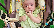 Olive is registered to the contest to win money with this photo: baby, infant, child, car_seat, stroller, portrait, sitting, wide_eyed, finger_sucking, headband, bow, green_dress, socks, toys, plush_toy, floral_pattern, indoor, green_background, hand, safety_labels