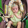 baby, infant, child, car_seat, stroller, portrait, sitting, wide_eyed, finger_sucking, headband, bow, green_dress, socks, toys, plush_toy, floral_pattern, indoor, green_background, hand, safety_labels