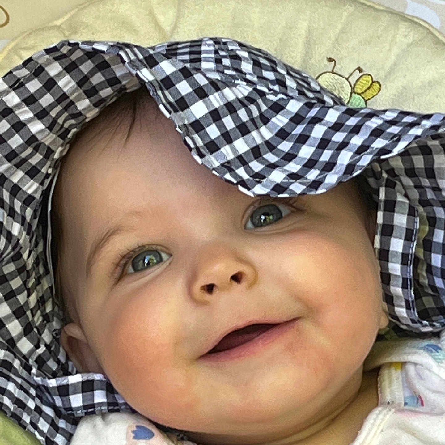 Zaiah is registered to the contest to win money with this photo: baby, baby_girl, bee_design, checkered_pattern, cheeks, child, close_up, clothing, cushion, cute, eyes, face, happy, hat, headwear, infant, lying_down, pattern, portrait, smiling