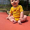 baby, barefoot, child, curious_expression, cute, daylight, feet, fence, grass, hands, head, infant, nature, outdoor, portrait, red_surface, sitting, smiling, sunny, yellow_onesie