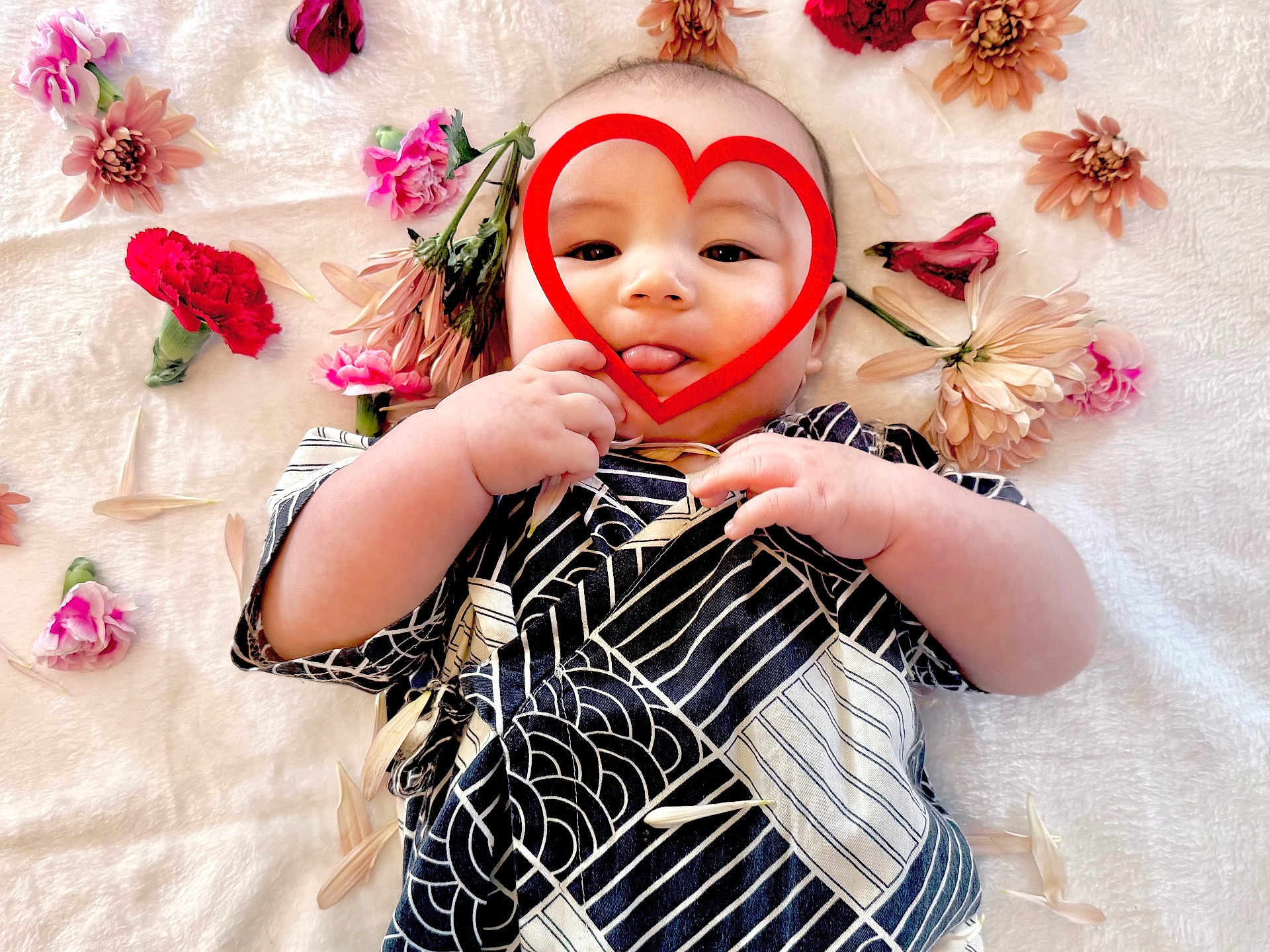 Zen is registered to the contest to win money with this photo: baby, infant, heart_shape, flowers, pink_flowers, red_flowers, blanket, patterned_clothing, tongue_out, cute, playful, soft_texture, lying_down, hand, face, portrait, child, adorable, decorative, peaceful