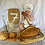 baby, chef_hat, wooden_spoon, bread, pastries, cutting_board, flour, whisk, measuring_cup, apron, smiling, sitting, white_background, kitchen_theme, child, baking, happy, portrait, indoor, food