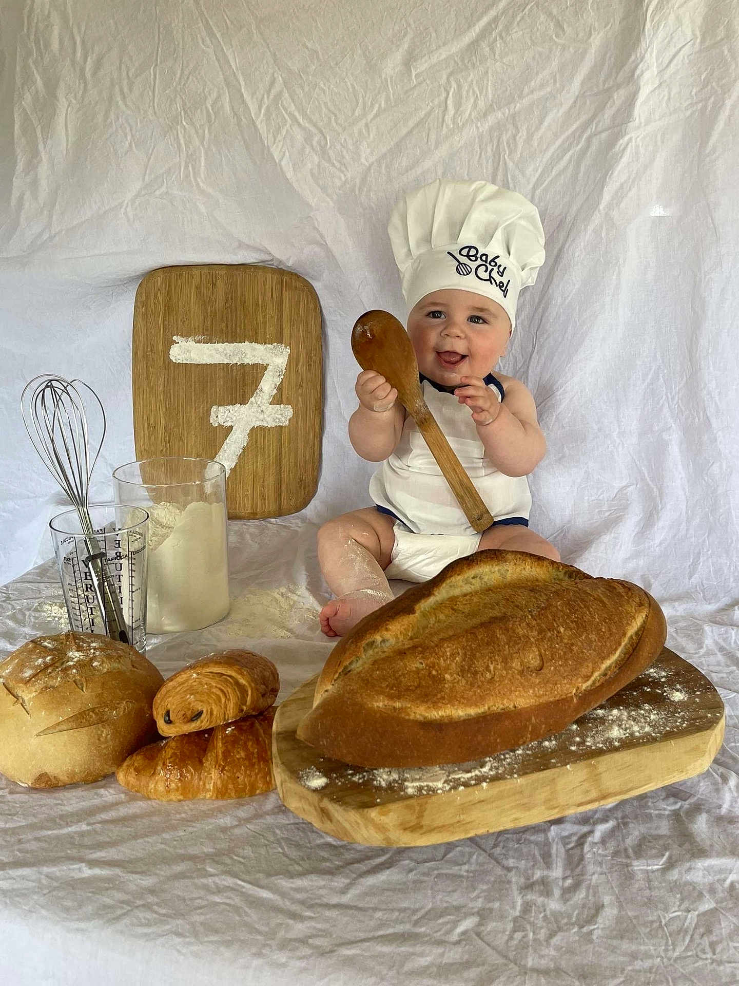 Akis participe au concours pour gagner de l'argent avec cette photo : baby, chef_hat, wooden_spoon, bread, pastries, cutting_board, flour, whisk, measuring_cup, apron, smiling, sitting, white_background, kitchen_theme, child, baking, happy, portrait, indoor, food