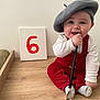 baby, beret, paintbrush, red_overalls, white_shirt, canvas, number_six, wooden_surface, sitting, happy, smiling, face, chewing, milestone, indoor, cute, child, person, sock, celebration