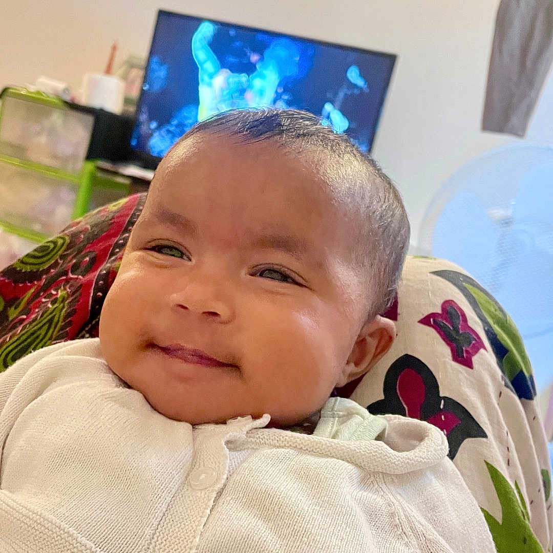 Tiana Talukder participe au concours pour gagner de l'argent avec cette photo : baby, blurred_background, child, closeup, clothing, comfort, cozy, cute, face, happy, head, indoor, infant, patterned_fabric, person, portrait, relaxing, smiling, television, white_cardigan