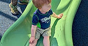 Elijah is registered to the contest to win money with this photo: toddler, child, slide, playground, shoes, dinosaur_shirt, red_hair, shorts, socks, adult_legs, green_slide, blue_floor, sunlight, shadow, outdoor, play, recreation, person, casual_clothing, fun