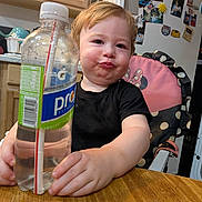 Elijah joined the competition — help win amazing prizes! toddler, child, water_bottle, high_chair, kitchen, fridge, magnets, photos, table, wood, black_shirt, pink_chair, polka_dots, face, expression, hand, cupboard, straw, drink, indoor