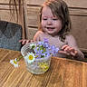 toddler, child, flowers, plastic_cup, table, wooden_table, kitchen, cabinet, chair, wildflowers, daisies, purple_flowers, yellow_flowers, curly_hair, person, indoor, smiling, young_child, casual, home