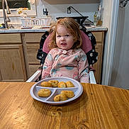 Willow joined the competition — help win amazing prizes! child, high_chair, plate, food, nuggets, table, wooden_table, pink_jacket, kitchen, cabinet, window, curtains, sink, dish_rack, person, indoor, meal, expression, young_child, cozy_clothing