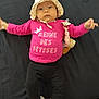 baby, infant, child, hat, pink_shirt, black_pants, pink_shoes, pacifier, stuffed_toy, lying_down, curious_expression, clothing, face, person, footwear, indoor, black_background, cute, portrait, young_child