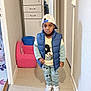 child, boy, indoor, hallway, clothing, cap, puffer_vest, mickey_mouse, sweatshirt, pants, sneakers, serious_expression, cabinet, chair, bucket, floor, wall, person, standing, portrait
