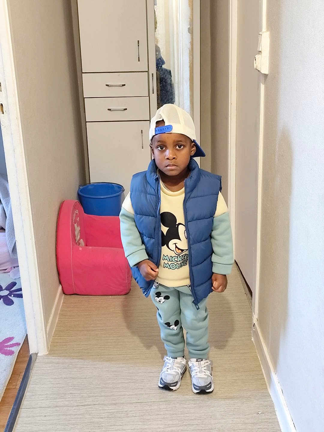 Ansry a rejoint le concours — aidez-le/la à gagner de superbes lots ! child, boy, indoor, hallway, clothing, cap, puffer_vest, mickey_mouse, sweatshirt, pants, sneakers, serious_expression, cabinet, chair, bucket, floor, wall, person, standing, portrait
