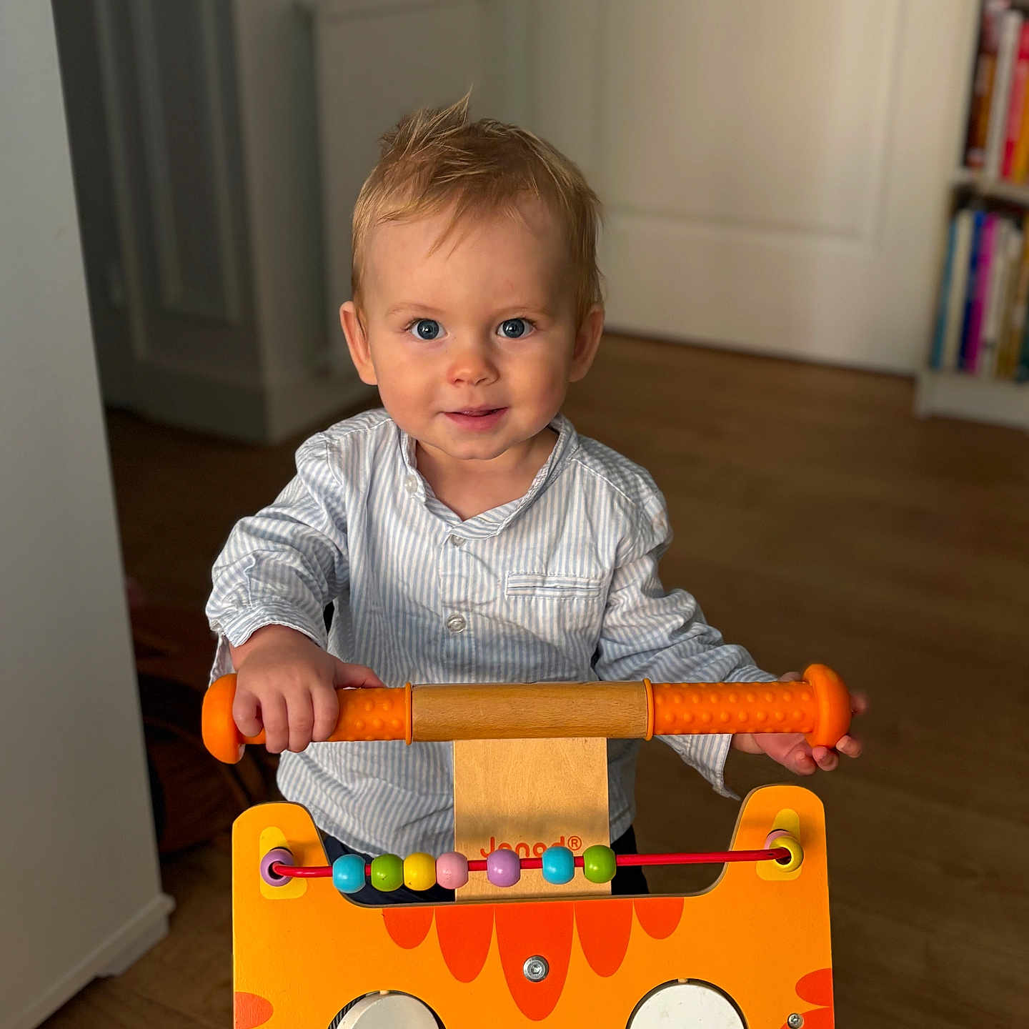 Henri participe au concours pour gagner de l'argent avec cette photo : toddler, child, toy_walker, orange_toy, cat_face, blue_eyes, indoor, wooden_floor, bookshelf, smiling, striped_shirt, handlebar, playroom, cute, person, floor, furniture, young_child, happy, home