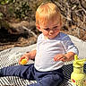 toddler, child, outdoor, blanket, food, snack, water_bottle, yellow, shoes, pants, shirt, nature, sunlight, expression, curious, sitting, striped, daylight, young_child, playful