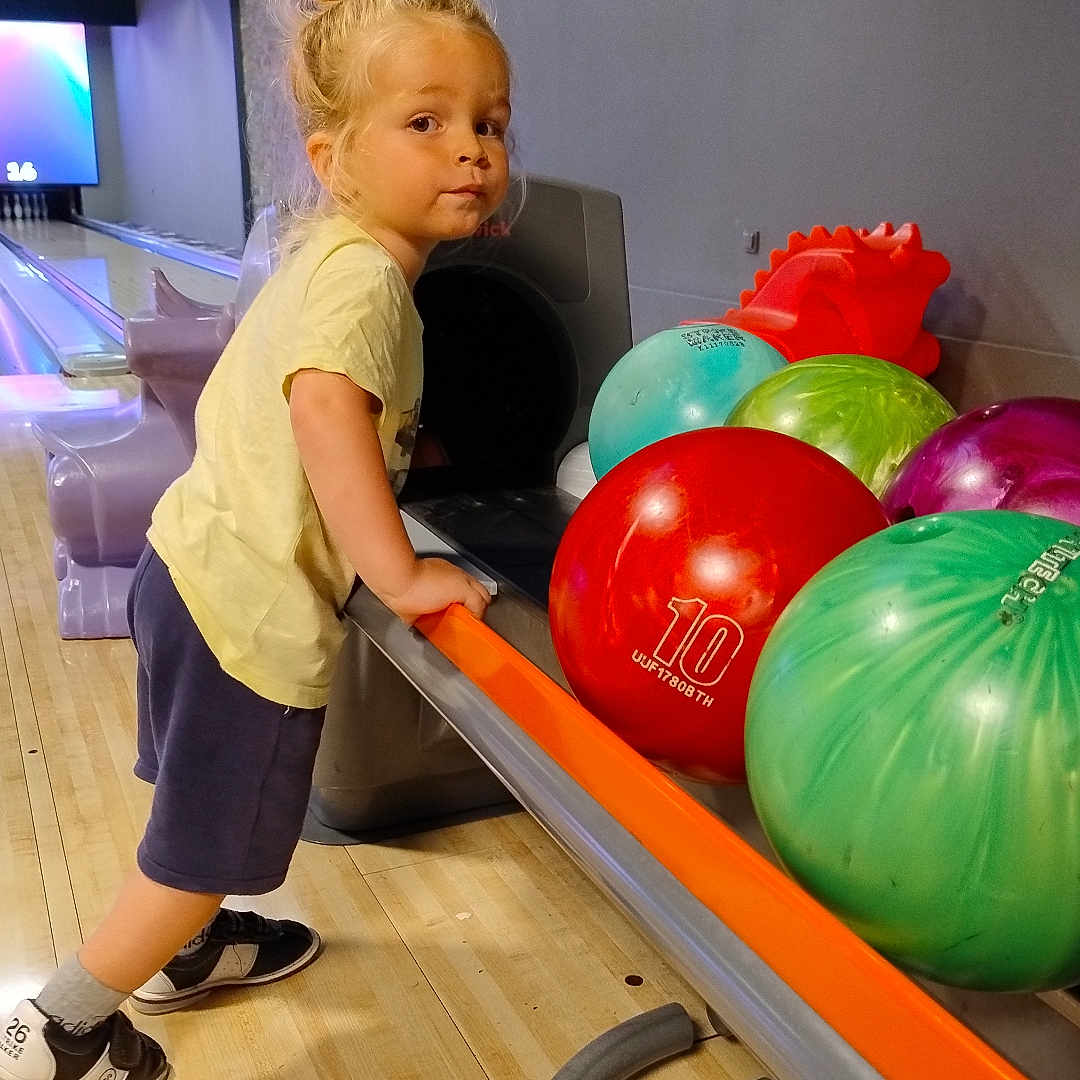 Loé participe au concours pour gagner de l'argent avec cette photo : activity, balls, blonde_hair, bowling_alley, bowling_balls, casual_clothing, child, colorful, curious, fun, indoor, leisure, person, rack, recreational, shoes, sports_equipment, standing, wooden_floor, young_child