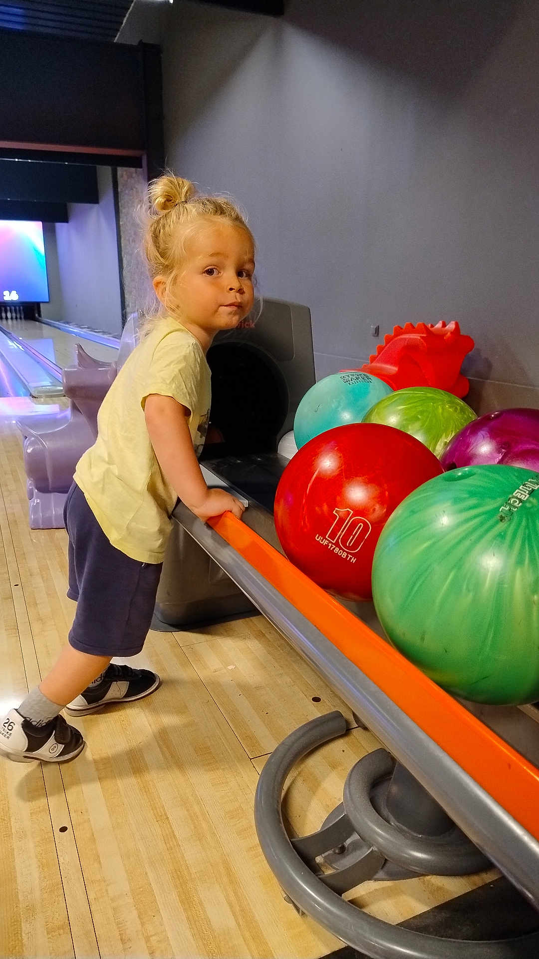 Loé participe au concours pour gagner de l'argent avec cette photo : child, bowling_alley, bowling_balls, curious, blonde_hair, casual_clothing, indoor, sports_equipment, leisure, young_child, shoes, wooden_floor, recreational, colorful, standing, person, activity, fun, balls, rack