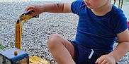 Loé participe au concours pour gagner de l'argent avec cette photo : toddler, child, toy, excavator, cat_brand, gravel, outdoor, braided_hair, blue_clothing, blue_shoes, play, nature, greenery, fence, sunny, summer, kid, construction_toy, cute, casual