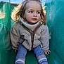 child, toddler, slide, playground, jacket, fleece, socks, shoes, outdoor, sunlight, tree, play, young_child, curly_hair, casual_clothing, park, fun, happy, closeup, daytime