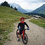 child, mountain_bike, helmet, protective_gear, gloves, outdoor, mountain, path, gravel, nature, greenery, hill, trees, cloudy_sky, rural, adventure, sport, youth, cycling, landscape