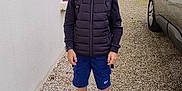 Elio participe au concours pour gagner de l'argent avec cette photo : child, boy, glasses, black_puffer_vest, blue_shorts, black_sneakers, gravel_path, house_wall, flower_pot, green_plants, car, outdoor, daylight, smiling, person, backpack, short_hair, casual_clothing, nature, sidewalk