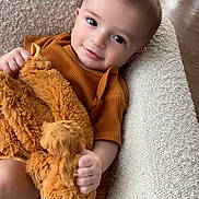 Cooper joined the competition — help win amazing prizes! baby, infant, child, smiling, plush_toy, stuffed_animal, couch, armchair, textured_fabric, brown_clothing, cozy, portrait, indoor, hand, eyes, hair, flooring, blanket, cute, relaxed