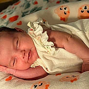 Monica is registered to the contest to win money with this photo: newborn, baby, sleeping, infant, portrait, face, indoors, blanket, bed, clothing, cream_outfit, peaceful, resting, curled_up, patterned_fabric, warm_lighting, close_up, candid, family_photo, adorable