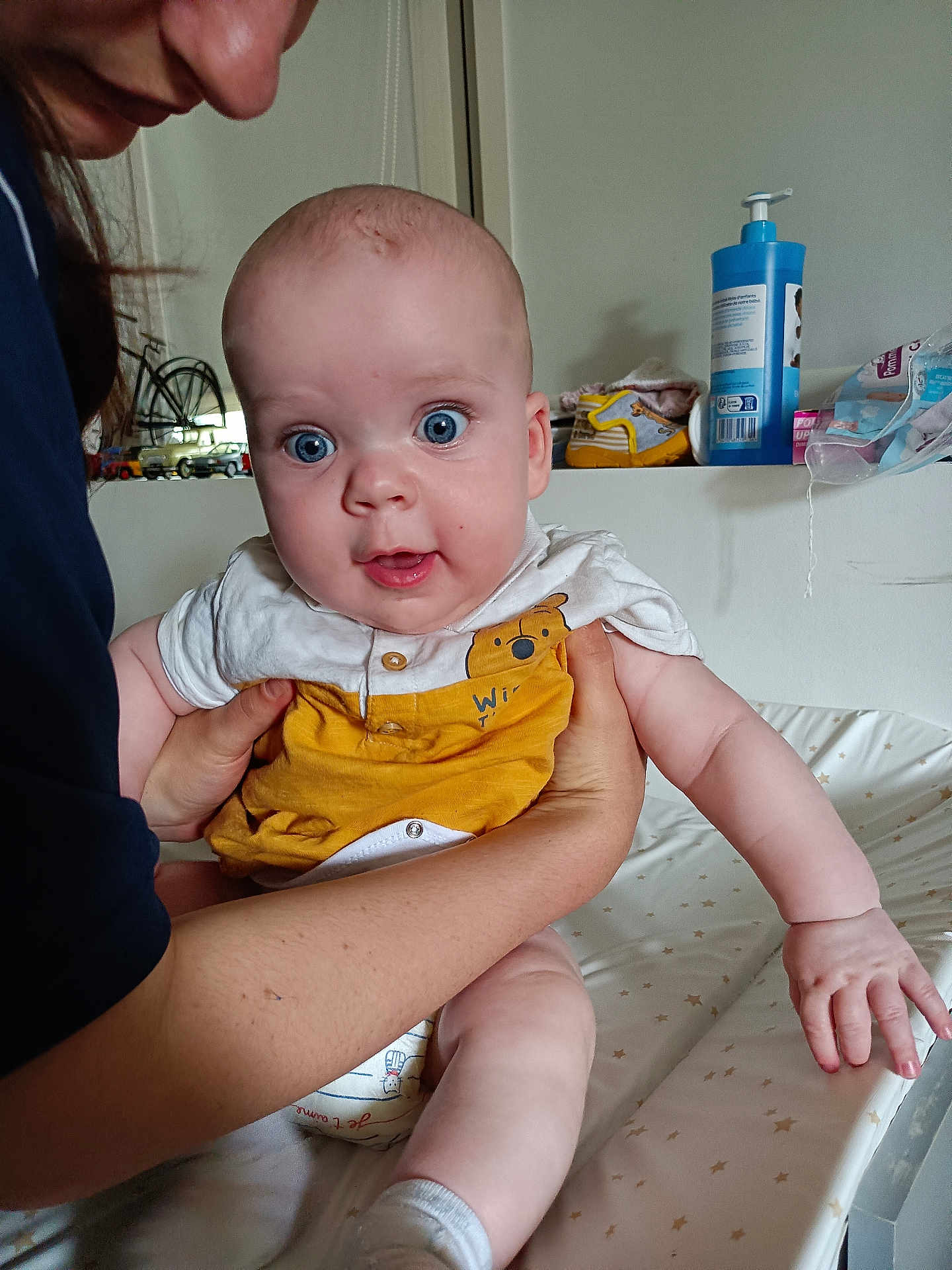 Alexandre a rejoint le concours — aidez-le/la à gagner de superbes lots ! baby, adult, blue_eyes, yellow_clothing, white_clothing, hands, infant, person, indoor, furniture, baby_changing_station, toy, skin, face, expression, holding, room, care_product, smile, curious