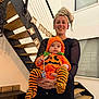 Louis a rejoint le concours — aidez-le/la à gagner de superbes lots ! baby, banner, child, costume, decor, festive, halloween, holding, home, indoor, modern, orange, person, pumpkin_costume, smile, staircase, striped_leggings, towel_headwrap, window, woman