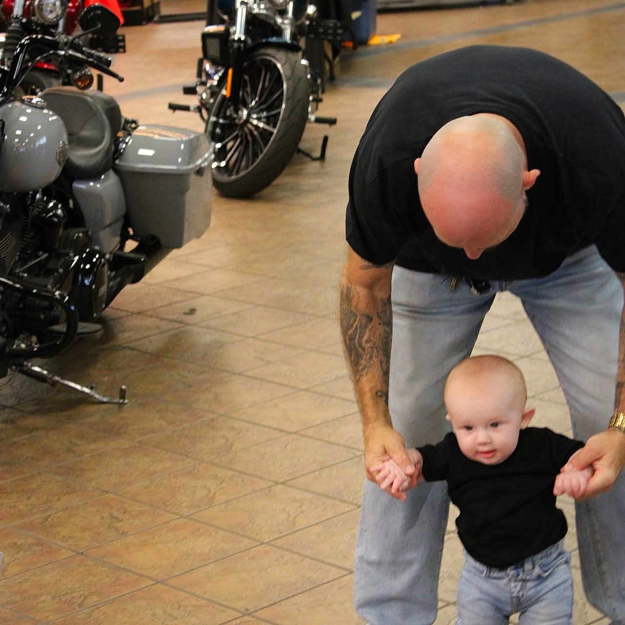 Jaxon is registered to the contest to win money with this photo: baby, child, man, tattoo, motorcycle, motorbike, showroom, floor, walking, supporting, indoor, person, head, black_shirt, jeans, footwear, people, shop, retail, helping